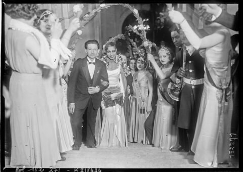 Joséphine Baker en robe blanche au bras de M. Poirier, sous une haie de rameaux fleuris, à la mairie du 20e arrondissement le 28 février 1931