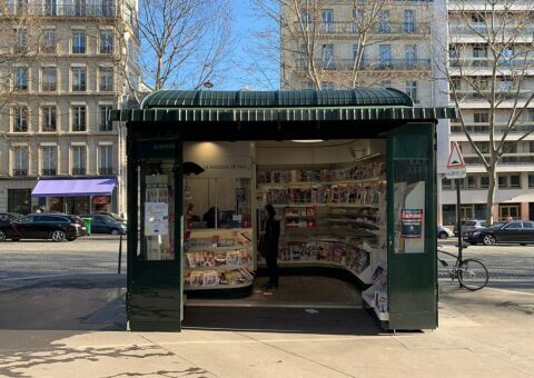 Un kiosque à journaux parisien vert, unes et magazines déployés sur l'avenue Marceau
