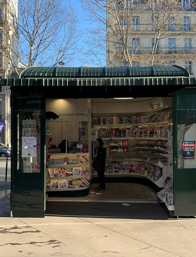 Un kiosque à journaux parisien vert, unes et magazines déployés sur l'avenue Marceau