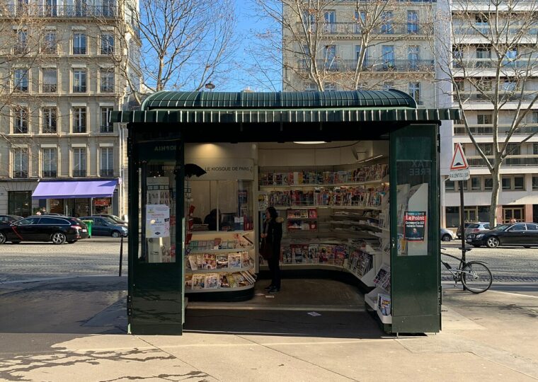 Un kiosque à journaux parisien vert, unes et magazines déployés sur l'avenue Marceau