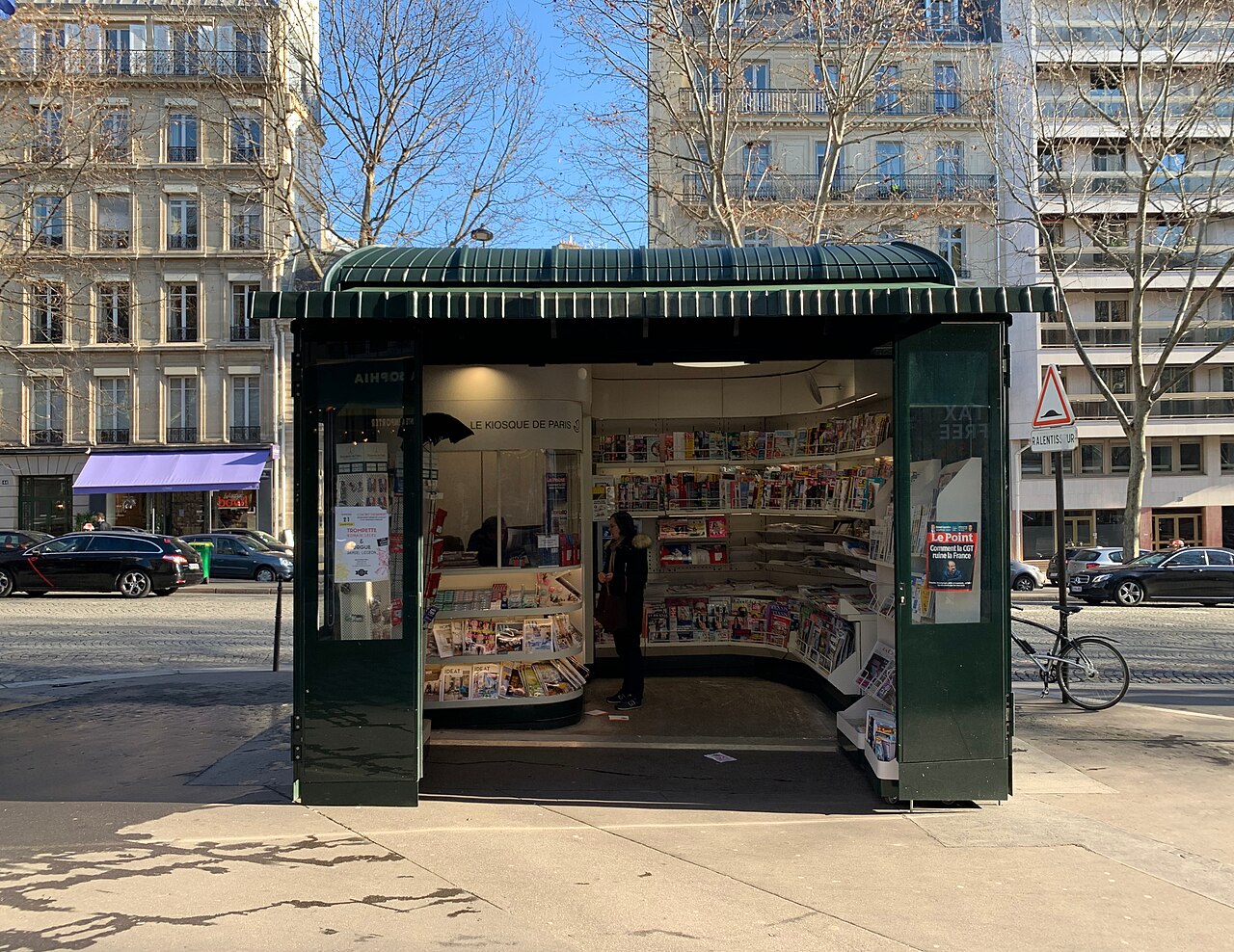 Un kiosque à journaux parisien vert, unes et magazines déployés sur l'avenue Marceau